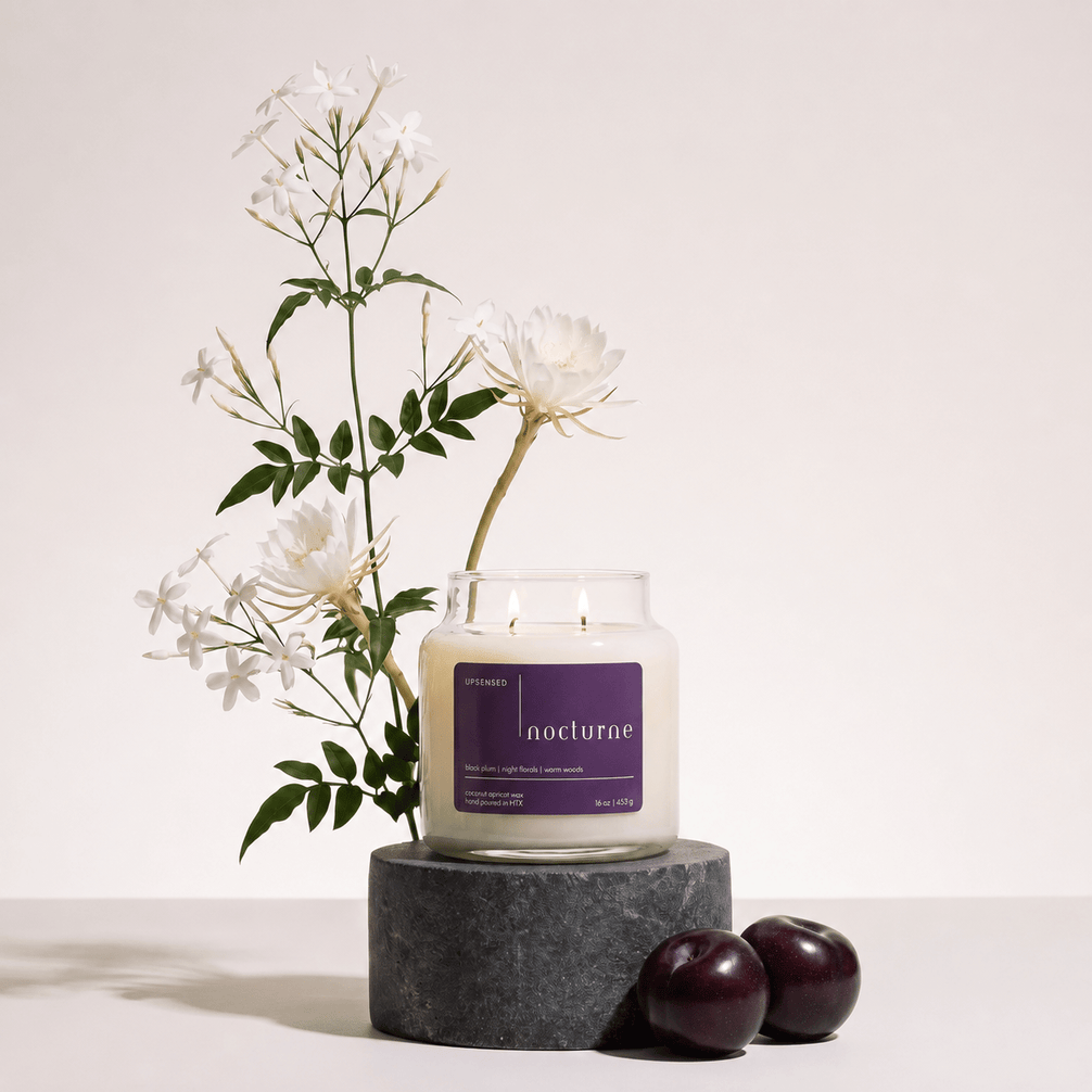 Upsensed Nocturne candle burning on a dark round stone pedestal, styled with white night-blooming jasmine and epiphyllum flowers and two dark plums.