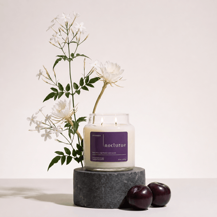 Upsensed Nocturne candle burning on a dark round stone pedestal, styled with white night-blooming jasmine and epiphyllum flowers and two dark plums.