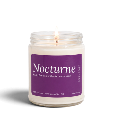 Upsensed Nocturne 8 oz soy candle, lit, in a clear glass jar with a plum purple label listing black plum, night florals, and warm woods