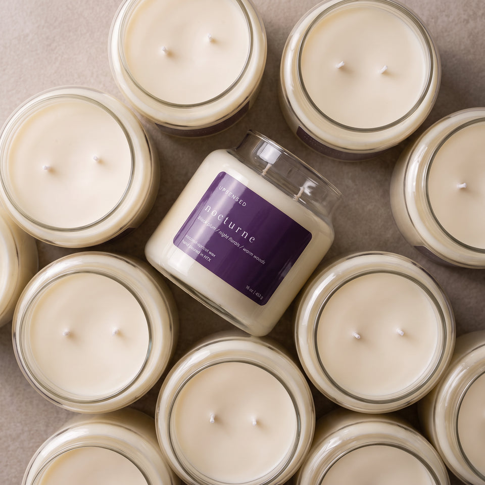 Overhead flat lay of Upsensed Nocturne candles in clear glass jars with creamy coconut-apricot wax and two white cotton wicks, featuring one candle on its side with a glossy deep plum label.