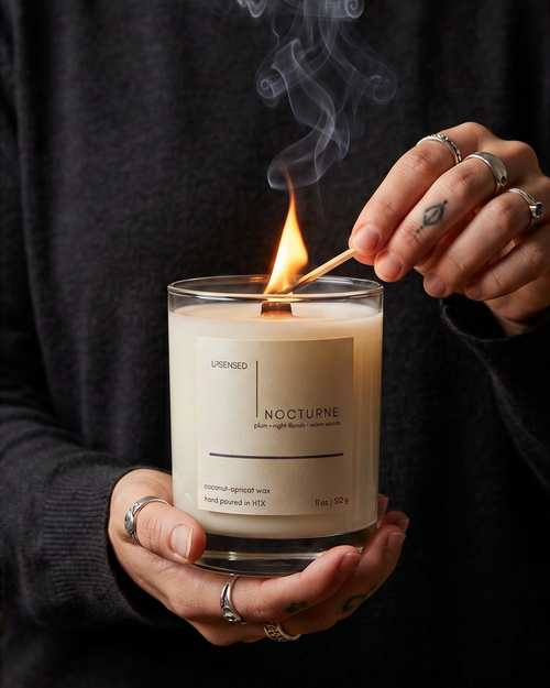 Person lighting an Upsensed Nocturne wooden wick candle with a match as soft smoke rises in a moody luxury home fragrance ritual.