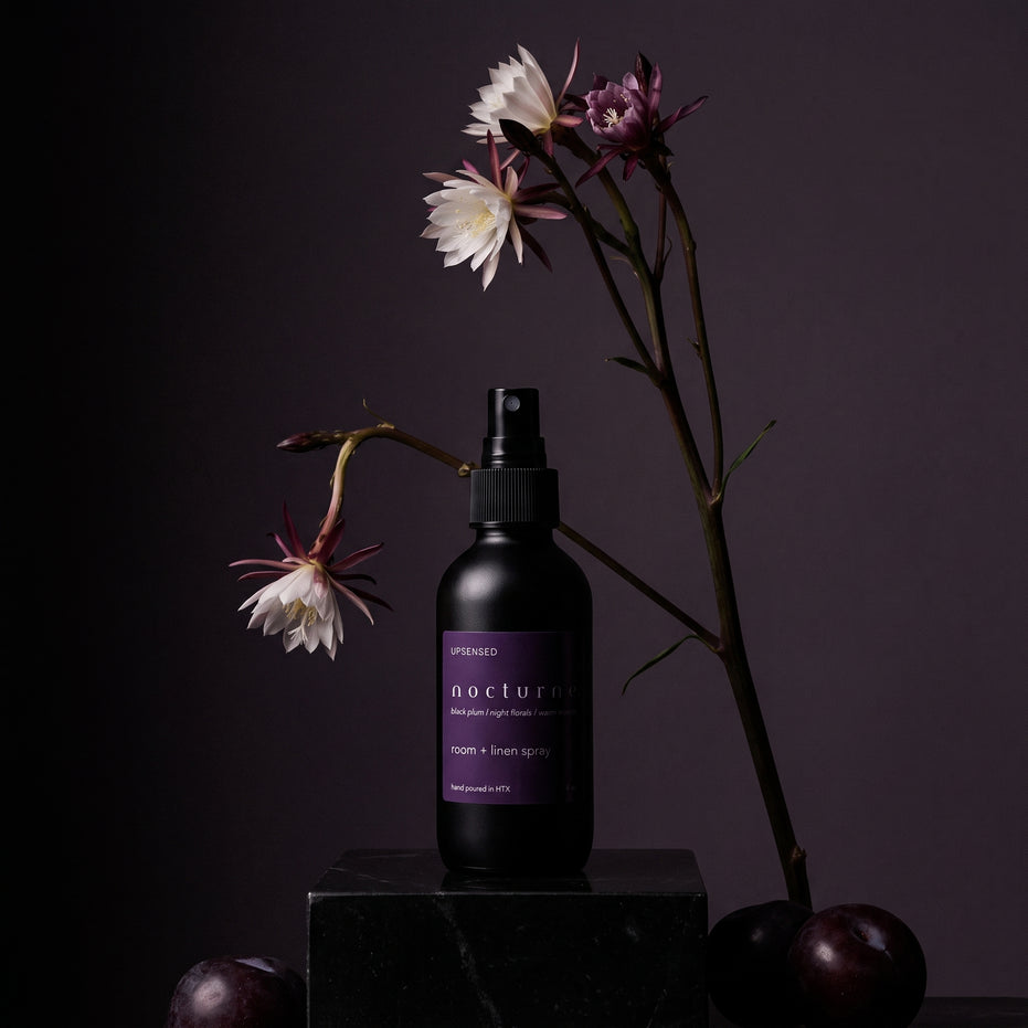 Nocturne room and linen spray on a dark plinth with night-blooming flowers and black plums.