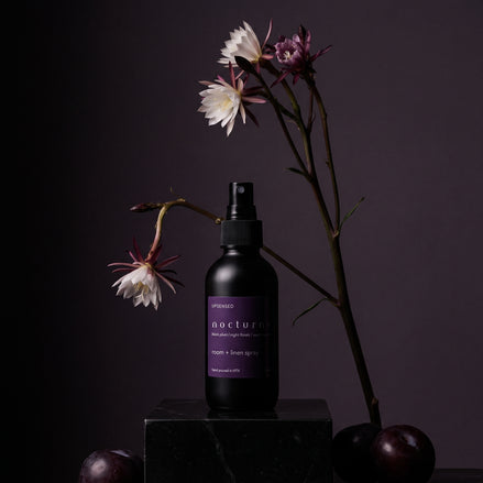 Nocturne room and linen spray on a dark plinth with night-blooming flowers and black plums.