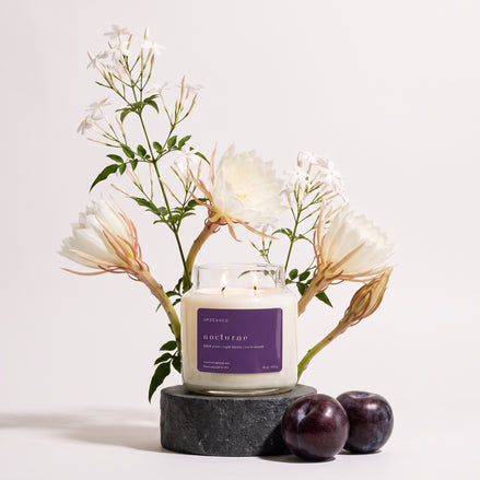 Upsensed Nocturne candle burning on a dark stone pedestal, styled with white night-blooming jasmine and epiphyllum flowers and two dark plums.