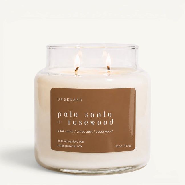 Upsensed Palo Santo + Rosewood 16 oz double-wick candle in a clear apothecary jar with a tan label, palo santo, citrus zest, and cedarwood scent notes, on a plain white background.