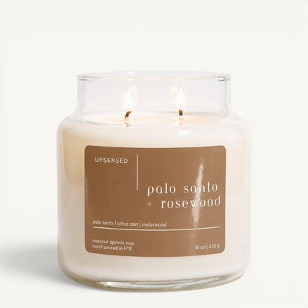 Upsensed Palo Santo + Rosewood 16 oz double-wick candle in a clear apothecary jar with a tan label, palo santo, citrus zest, and cedarwood scent notes, on a plain white background.