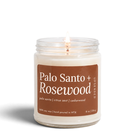 Upsensed Palo Santo + Rosewood 8 oz soy candle, lit, in a clear glass jar with a terracotta label listing palo santo, citrus zest, and cedarwood