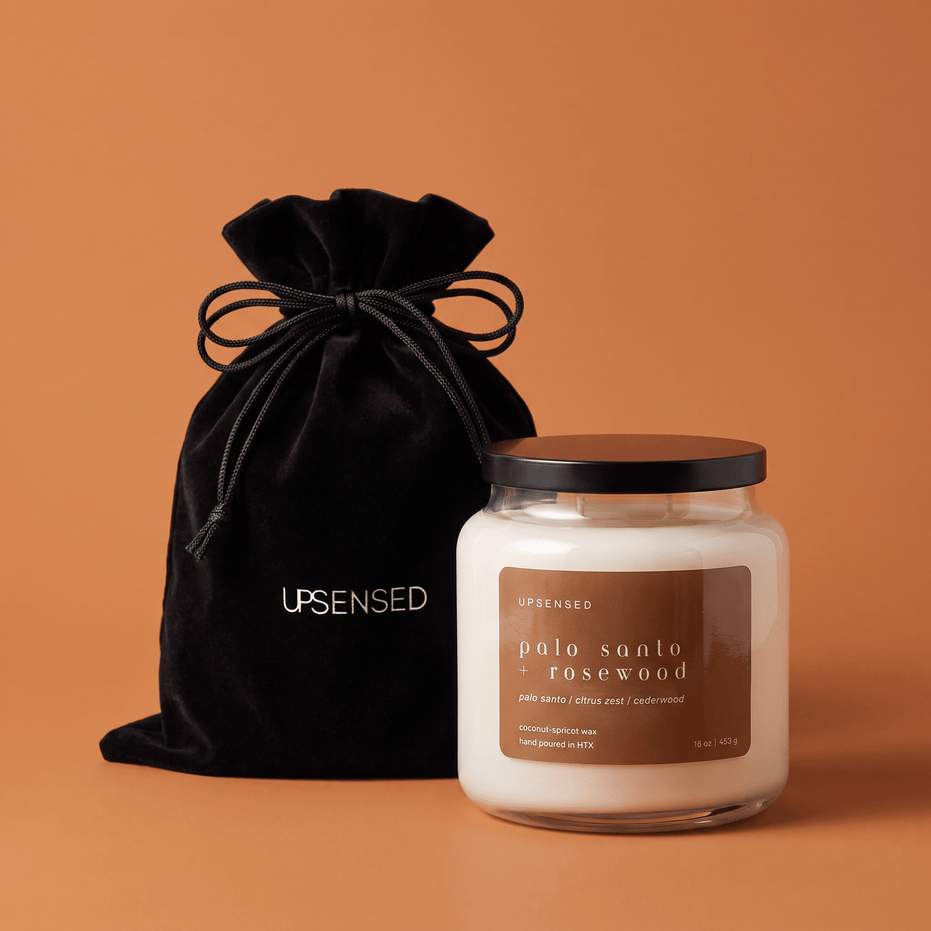 Upsensed Palo Santo + Rosewood candle with a black lid beside its black velvet UPSENSED drawstring packaging bag, on a burnt orange background.