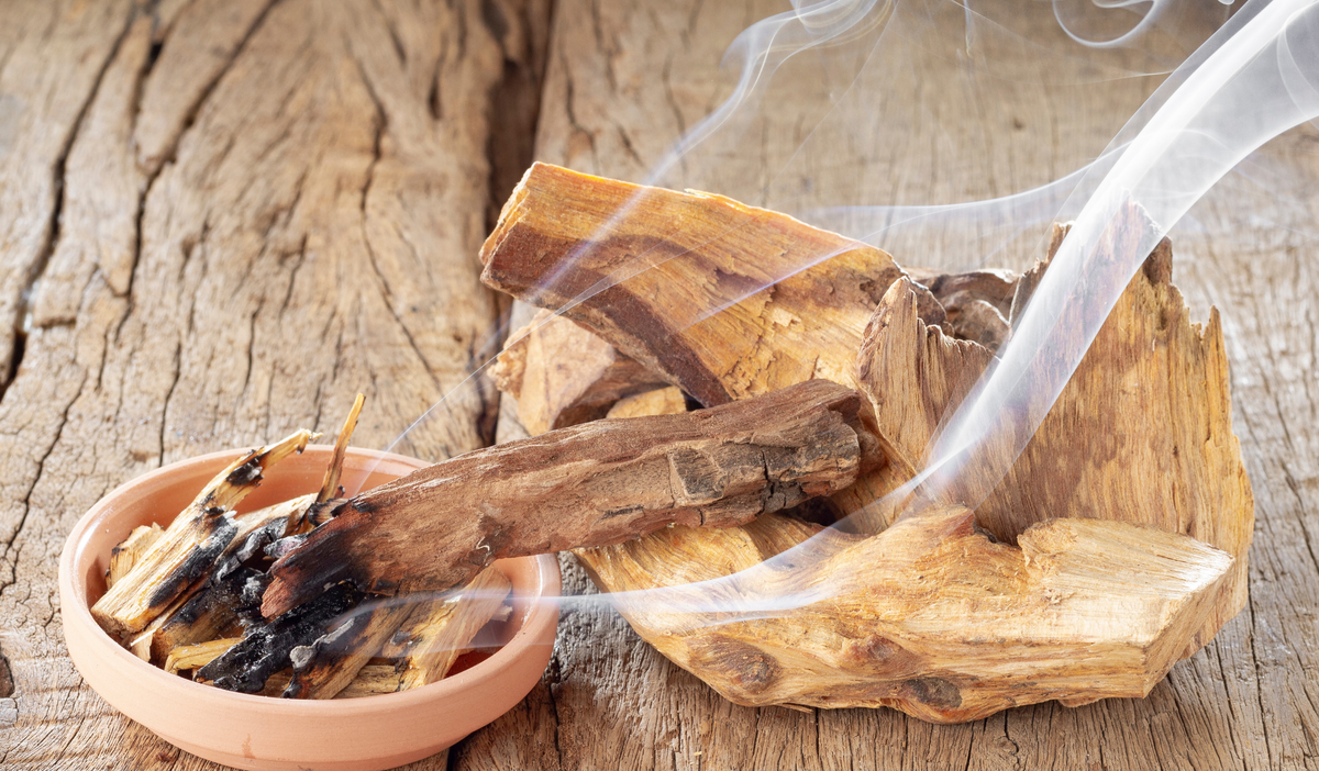 A small terracotta bowl filled with dried wood pieces and a lit smoldering piece, with visible smoke rising over stacked natural wood logs on a rustic wooden surface.