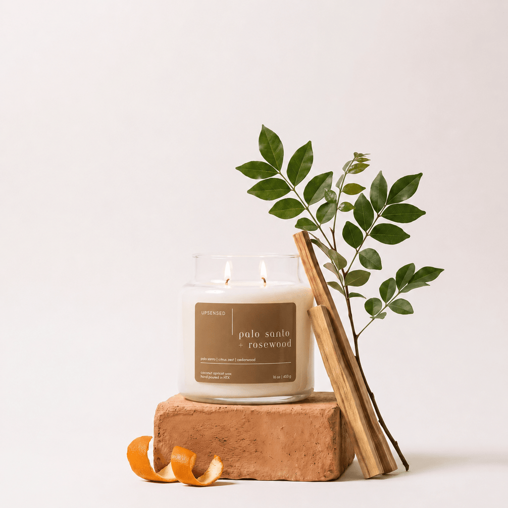 Upsensed Palo Santo + Rosewood candle burning on a terracotta brick, styled with a leafy branch, palo santo sticks, and a curled orange peel against a neutral background.