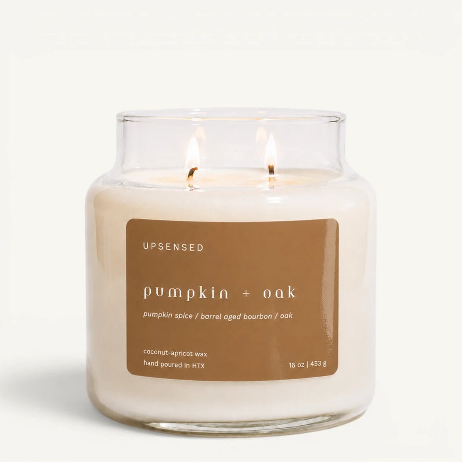 An Upsensed 'pumpkin + oak' scented candle in a clear glass jar with two lit wicks, featuring a brown label on a clean white background.