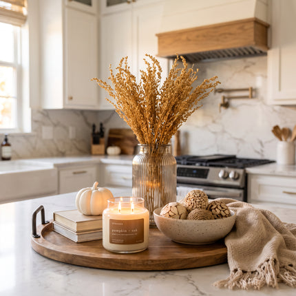 A lit candle in a glass jar sits on a wooden tray beside a small pumpkin, stacked books, a bowl of decorative items, and a vase of dried grasses, set on a marble kitchen counter.