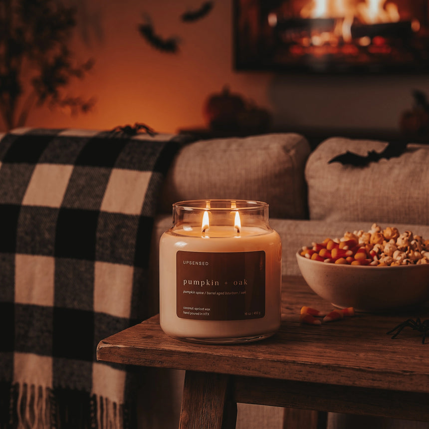 A lit candle in a clear glass jar sits on a wooden table in a cozy living room setting, with a black-and-white plaid throw blanket, a bowl of popcorn, and a warm fireplace in the background.