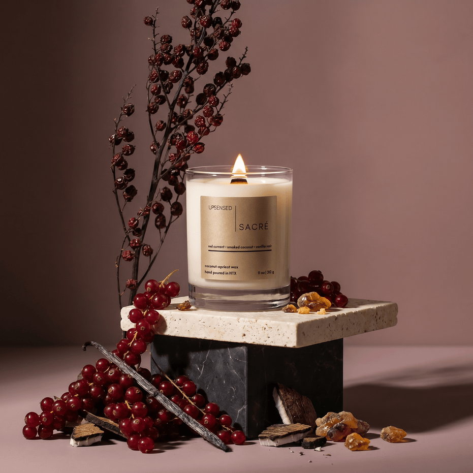Upsensed Sacré candle burning on a stacked stone and marble pedestal surrounded by red currant branches, vanilla beans, smoked coconut pieces, and amber resin against a warm mauve background