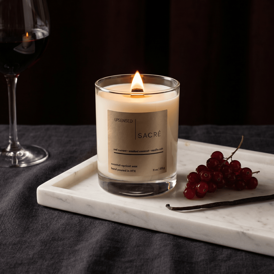Upsensed Sacré candle burning on a marble tray beside fresh red currants, a vanilla bean, and a glass of red wine against a dark moody background