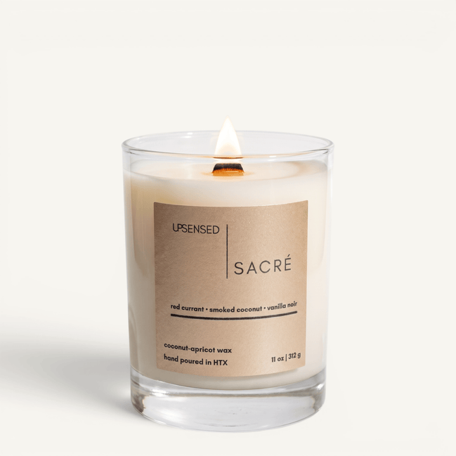 Upsensed Sacré candle lit on white background, showing warm gold label with red currant, smoked coconut, and vanilla noir notes