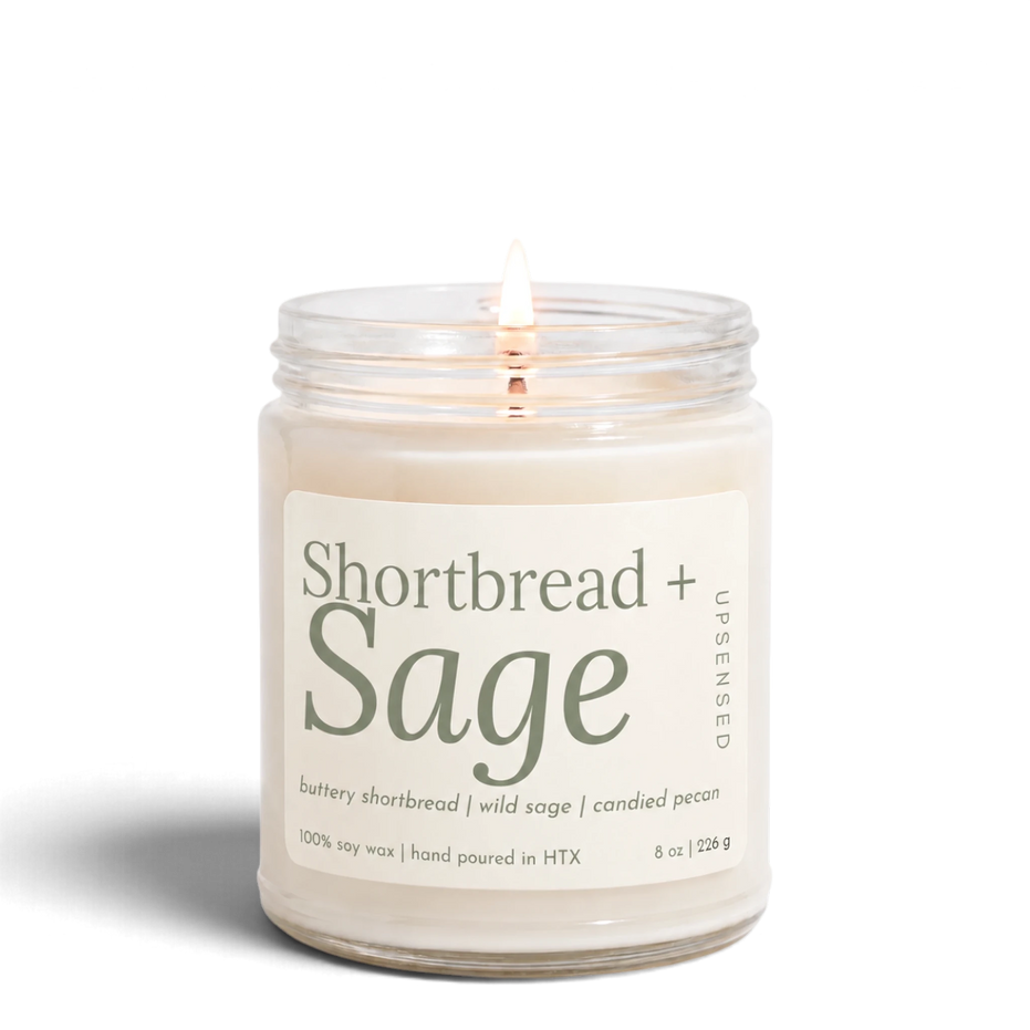 Upsensed Shortbread + Sage 8 oz soy candle, lit, in a clear glass jar with a cream label and sage green lettering listing buttery shortbread, wild sage, and candied pecan