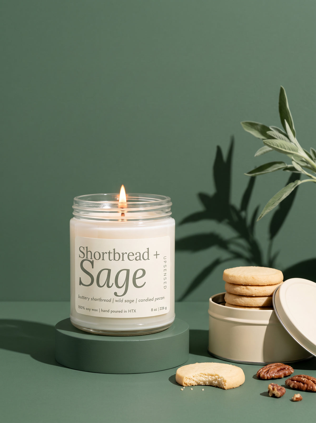 A lit candle in a clear glass jar sits on a round pedestal against a muted green background. The label reads 'Shortbread + Sage'