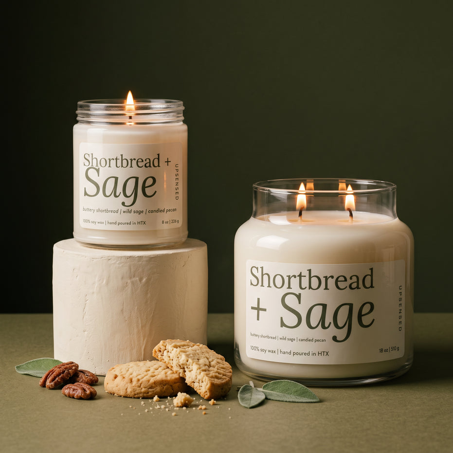 Shortbread + Sage soy candle in two sizes: a lit 8 oz single-wick jar on a cream plaster pedestal beside a lit 18 oz double-wick jar, both with cream labels, with shortbread cookies, candied pecans, and sage leaves against an olive green background