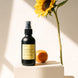 Sunflowers + Citrus room and linen spray on a cream pedestal beside a mandarin orange and sunflower.