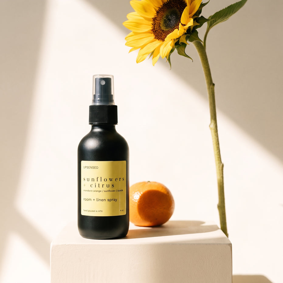 Sunflowers + Citrus room and linen spray on a cream pedestal beside a mandarin orange and sunflower.
