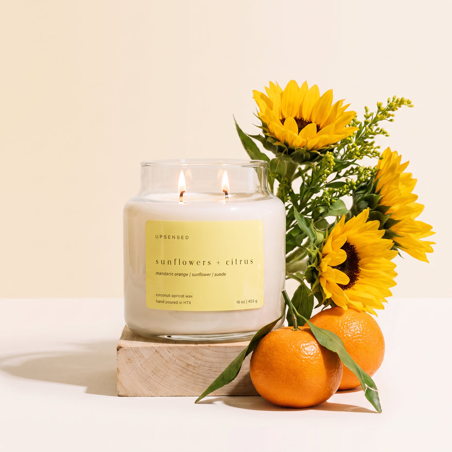 Upsensed Sunflowers + Citrus candle on a light wood block, styled with fresh sunflowers and mandarin oranges against a cream background.