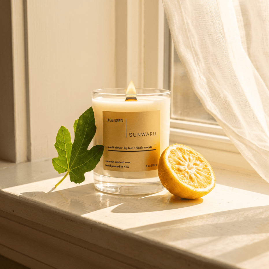 Upsensed Sunward citrus wood candle burning on a sunlit windowsill beside a fresh fig leaf and halved lemon