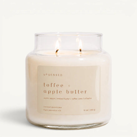 A lit two-wick candle in a clear glass jar with cream-colored wax and a beige label reading 'UPSENSED toffee + apple butter' on a clean white background.