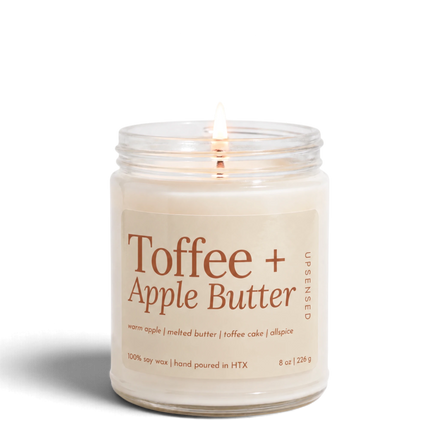 Upsensed Toffee + Apple Butter 8 oz soy candle, lit, in a clear glass jar with a sand-colored label and copper lettering listing warm apple, melted butter, toffee cake, and allspice