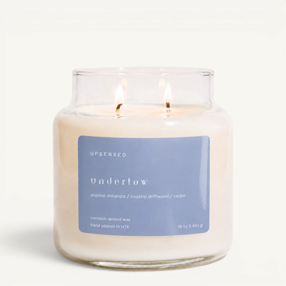 Upsensed Undertow 16 oz double-wick candle in a clear apothecary jar with a dusty blue label, marine minerals, coastal driftwood, and cedar scent notes, on a plain white background.
