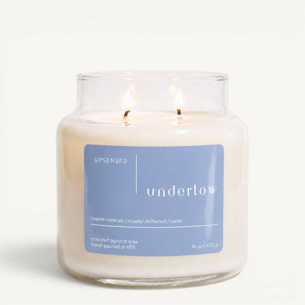 Upsensed Undertow 16 oz double-wick candle in a clear apothecary jar with a dusty blue label, marine minerals, coastal driftwood, and cedar scent notes, on a plain white background.