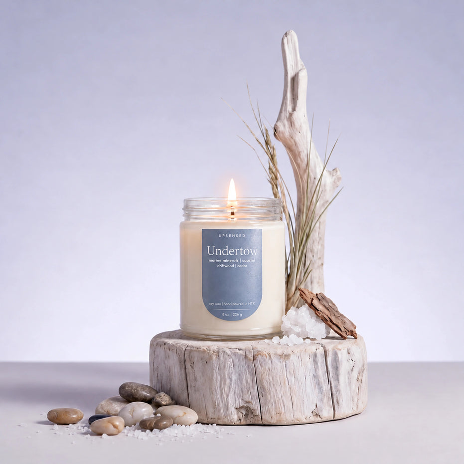 Upsensed Undertow 8 oz single-wick candle lit on a whitewashed driftwood round with sea salt, beach stones and dune grass