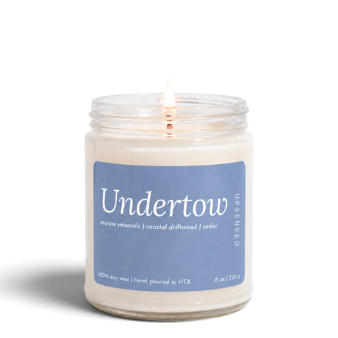 Upsensed Undertow 8 oz soy candle, lit, in a clear glass jar with a dusty blue label listing marine minerals, coastal driftwood, and cedar