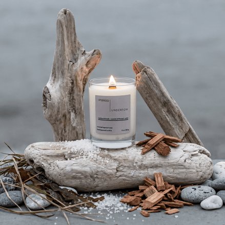 Upsensed Undertow candle burning on a large driftwood log at the coast, surrounded by sea salt, cedar wood chips, and smooth river stones