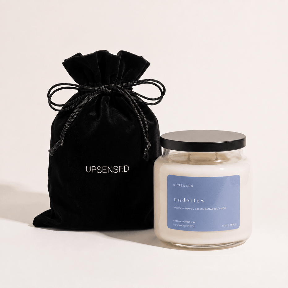 Upsensed Undertow candle with a black lid beside its black velvet UPSENSED drawstring packaging bag, on a cream background.