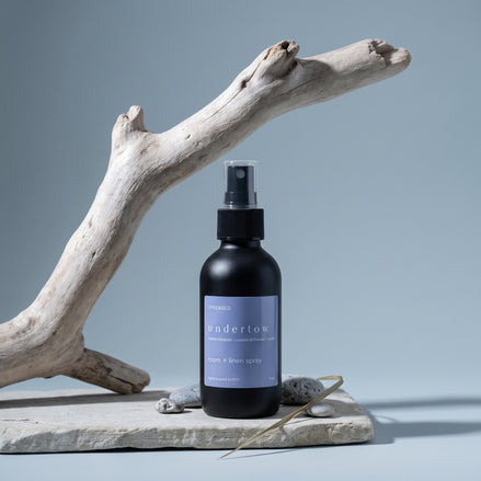 Undertow room and linen spray on a stone slab with driftwood, grey pebbles, and dried sea grass.