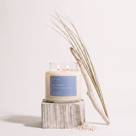 Upsensed Undertow candle burning on a weathered wood block, styled with a piece of driftwood, dried coastal grass, and scattered sea salt.