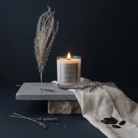 Upsensed Veil candle burning on a stacked stone pedestal surrounded by dried lavender stems, tonka beans, linen fabric, and pampas grass against a deep navy background