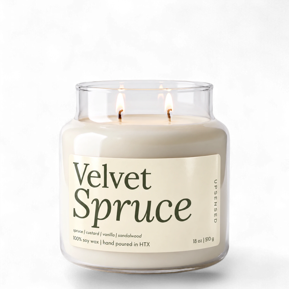 Upsensed Velvet Spruce 18 oz double-wick soy candle, lit, in a clear apothecary jar with an ivory label and deep green lettering listing spruce, custard, vanilla, and sandalwood