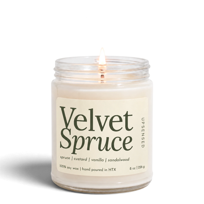 Upsensed Velvet Spruce 8 oz soy candle, lit, in a clear glass jar with an ivory label and deep green lettering listing spruce, custard, vanilla, and sandalwood
