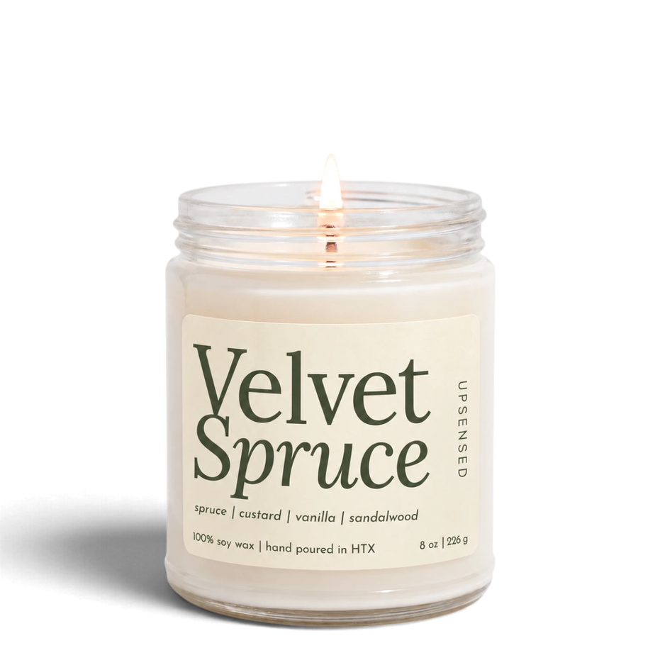 Upsensed Velvet Spruce 8 oz soy candle, lit, in a clear glass jar with an ivory label and deep green lettering listing spruce, custard, vanilla, and sandalwood