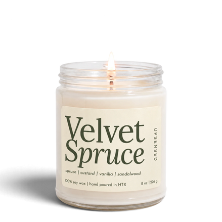 Upsensed Velvet Spruce 8 oz soy candle, lit, in a clear glass jar with an ivory label and deep green lettering listing spruce, custard, vanilla, and sandalwood