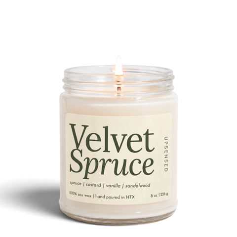 Upsensed Velvet Spruce 8 oz soy candle, lit, in a clear glass jar with an ivory label and deep green lettering listing spruce, custard, vanilla, and sandalwood