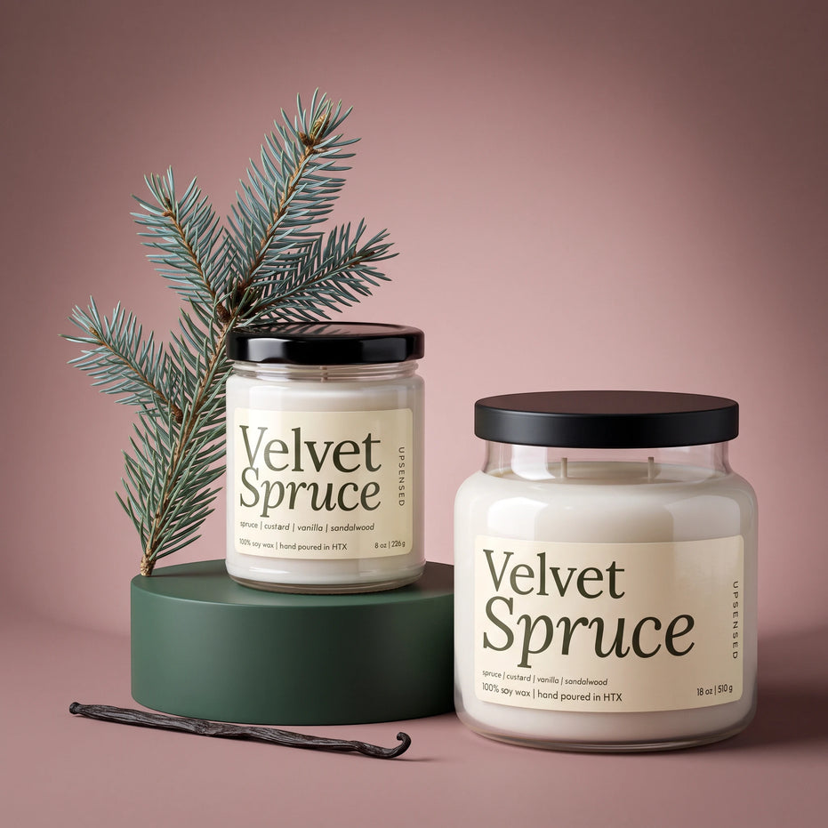 Two glass jar candles labeled 'Velvet Spruce' with black lids, styled on a dusty pink background with a green pedestal, pine branch, and vanilla bean.