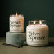 Velvet Spruce soy candle in two sizes: a lit 8 oz single-wick jar on a sage green pedestal beside a lit 18 oz double-wick jar, both with cream labels, with vanilla beans and a spruce sprig against a dark green background