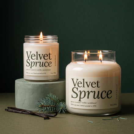 Velvet Spruce soy candle in two sizes: a lit 8 oz single-wick jar on a sage green pedestal beside a lit 18 oz double-wick jar, both with cream labels, with vanilla beans and a spruce sprig against a dark green background