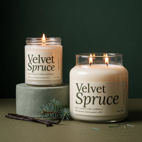 Velvet Spruce soy candle in two sizes: a lit 8 oz single-wick jar on a sage green pedestal beside a lit 18 oz double-wick jar, both with cream labels, with vanilla beans and a spruce sprig against a dark green background