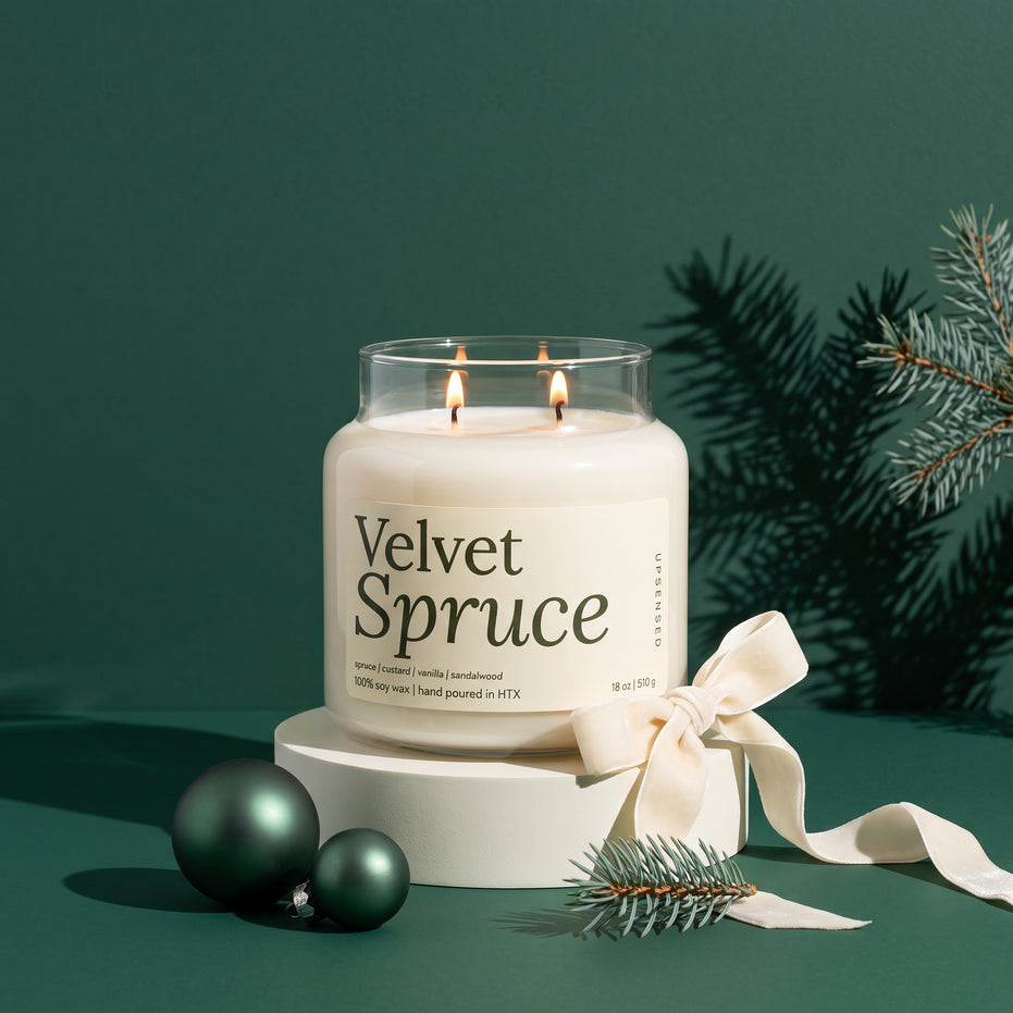 Velvet Spruce double-wick soy candle in an 18 oz glass jar with a cream label, lit on a cream pedestal with a cream velvet ribbon, green ornaments, and spruce branches against a dark green background
