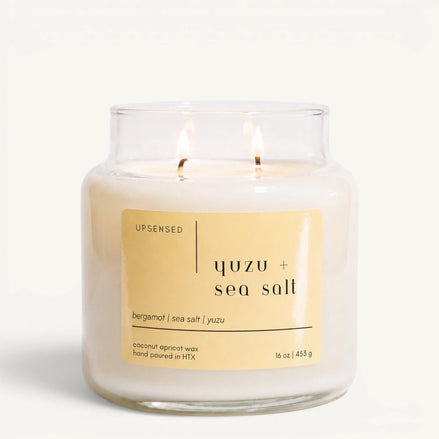 Upsensed Yuzu + Sea Salt 16 oz double-wick candle in a clear apothecary jar with a pale yellow label, bergamot, sea salt, and yuzu scent notes, on a plain white background.