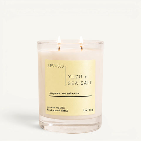 Yuzu + Sea Salt candle with gold label in a clear glass jar, two wicks lit.